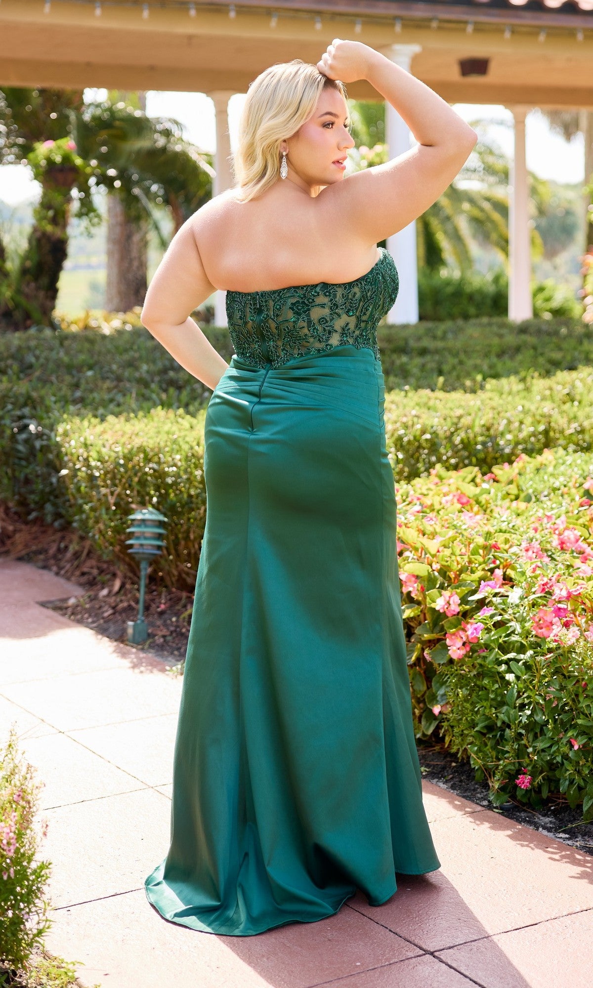A woman wears the Long Plus-Size Prom Dress Primavera 14109, featuring a strapless satin sheath skirt and sheer embroidered bodice, as she stands outdoors on a brick path amid greenery and pink flowers, her hand raised to her head.